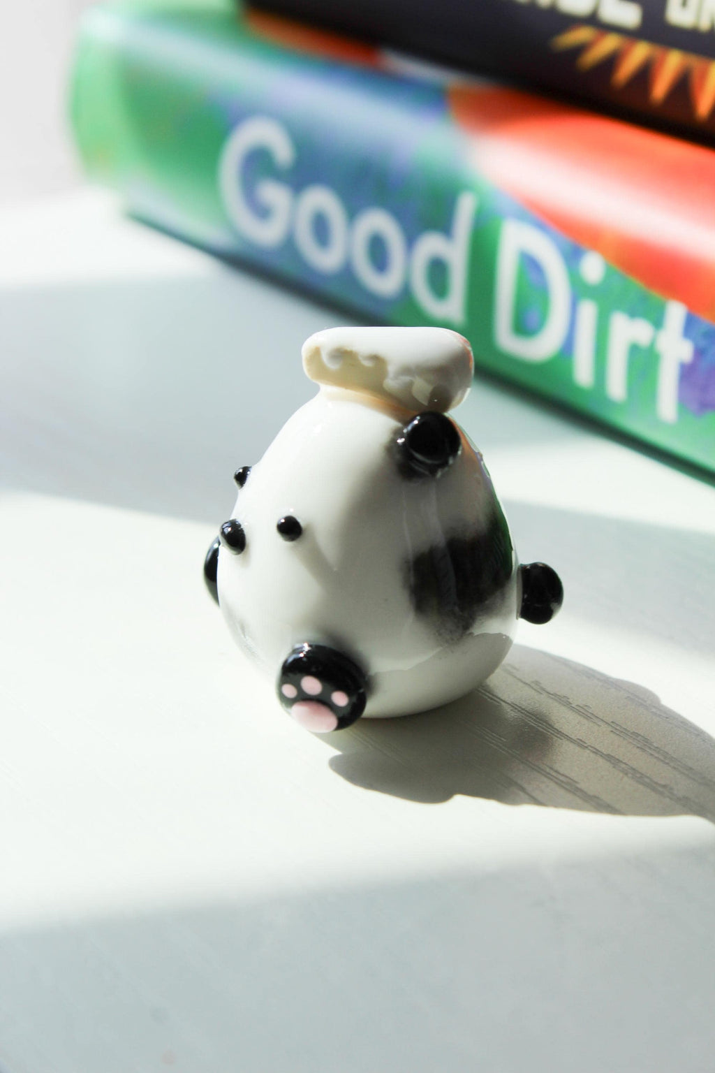 Small white figurine of a Panda with black accents and a piece of cake on a white surface, with a book titled 'Good Dirt' by Charmaine Wilkerson in the background.