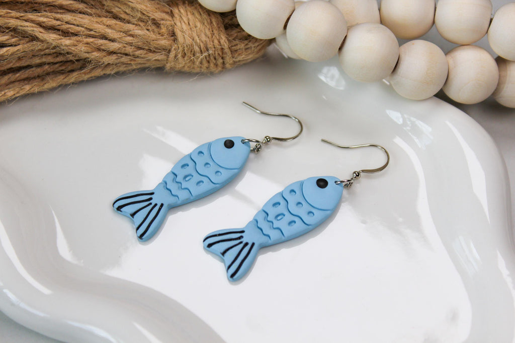 Blue fish-shaped earrings on a white dish with wooden beads in the background