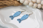 Blue fish-shaped earrings on a white dish with wooden beads in the background