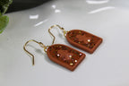 Pair of earrings with gold hooks, gingerbread men and terracotta-colored arch-shaped pendants on a white surface