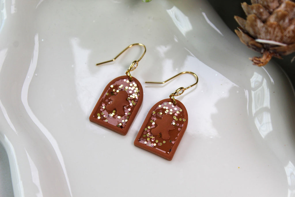 Pair of earrings with gold hooks, gingerbread men and terracotta-colored arch-shaped pendants on a white surface