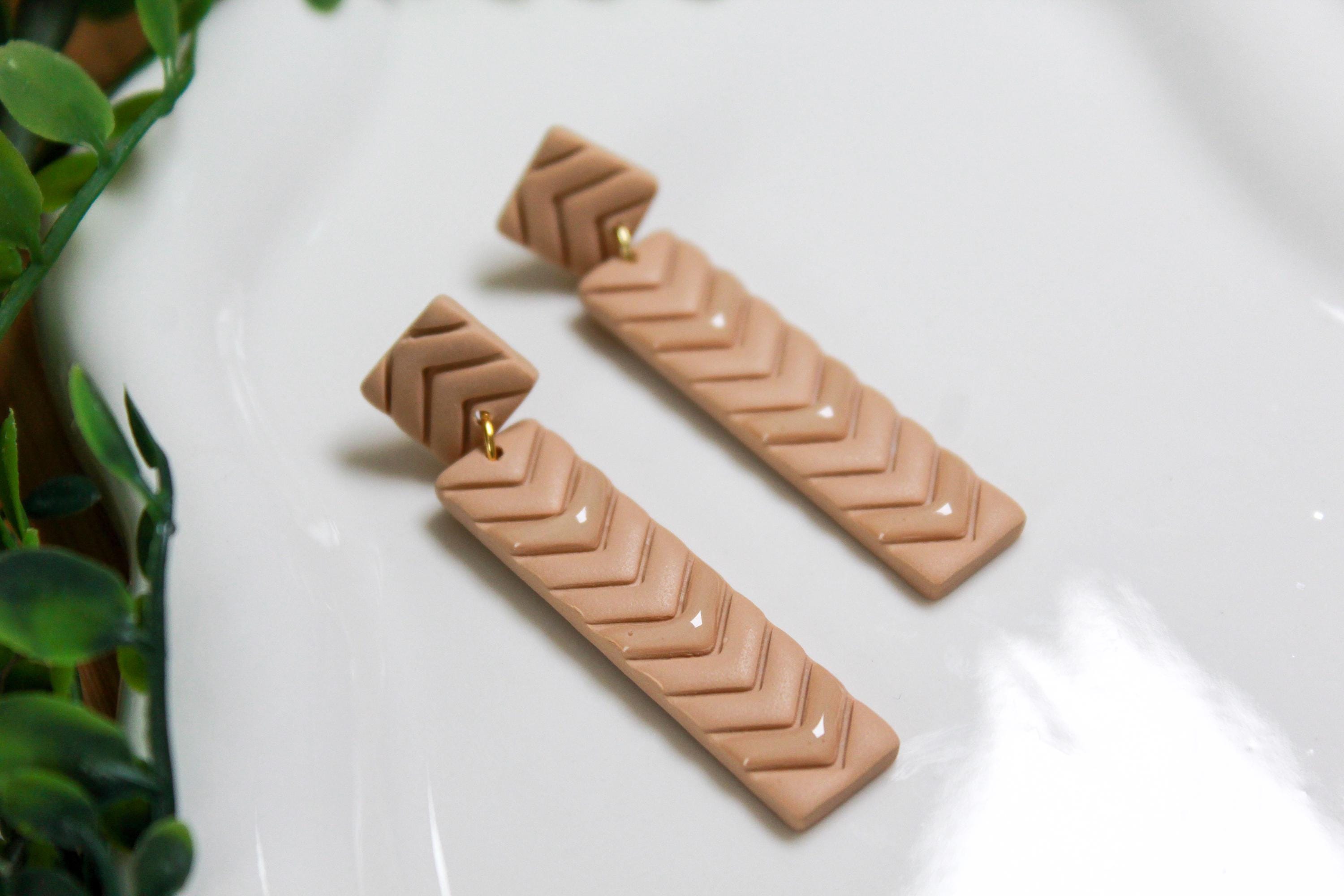 Pair of beige chevron patterned earrings on a white surface with green leaves in the background.