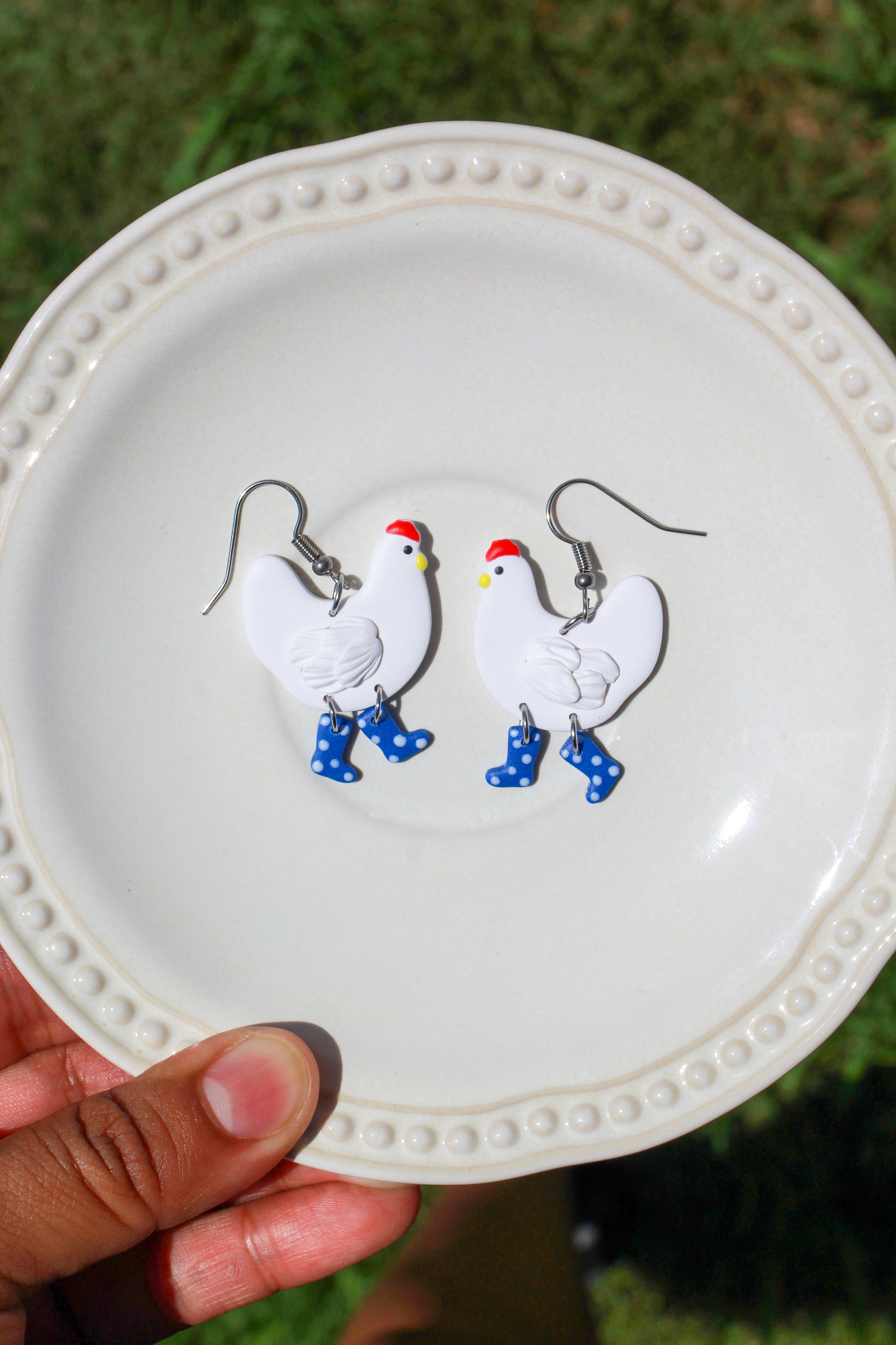 Chicken-shaped earrings with blue polkadot boots on a white plate held by a hand with a blurred green background