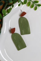 Green cactus-shaped earrings on a white plate with green leaves in the background