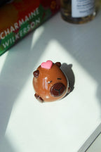 Small figurine of a Capybara with a pink heart on its head, placed on a surface with a book in the background.