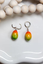 Pair of colorful mango-shaped earrings on a white surface with a blurred background.