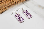 Pair of purple floral earrings on a white surface with dried grass in the background