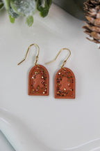 Pair of earrings with gold hooks, gingerbread men and terracotta-colored arch-shaped pendants on a white surface.