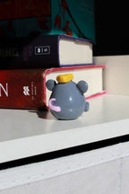 Small gray mouse figurine with a yellow hat and pink tongue on a shelf with books in the background