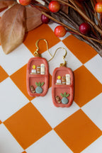 Pair of earrings resembling small shelves with miniature items on a checkered orange and white surface.