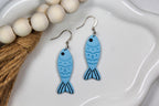 Blue fish-shaped earrings on a white dish with wooden beads and twine.