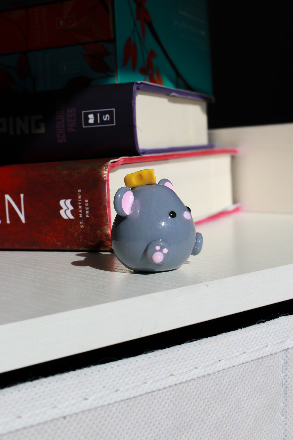 Small gray mouse figurine with pink accents on a shelf with books in the background
