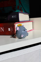 Small gray mouse figurine with pink accents on a shelf with books in the background