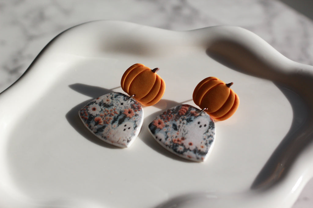Earrings with pumpkin-shaped tops and floral-patterned fields with ghosts at the bottom on a white surface