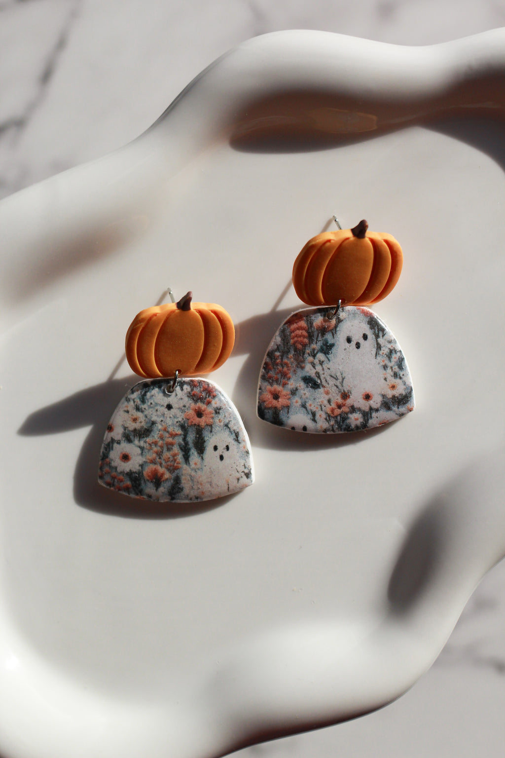 Earrings with pumpkin-shaped tops and floral-patterned fields with ghosts at the bottom on a white surface