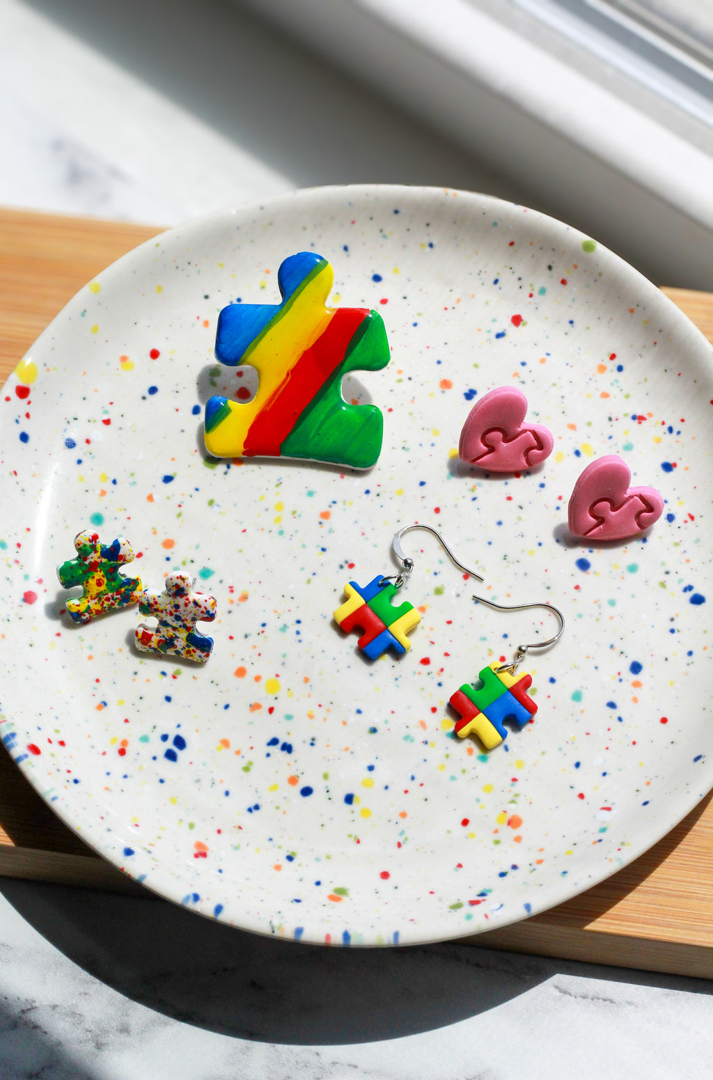 Colorful puzzle piece earrings on a speckled plate with a window in the background