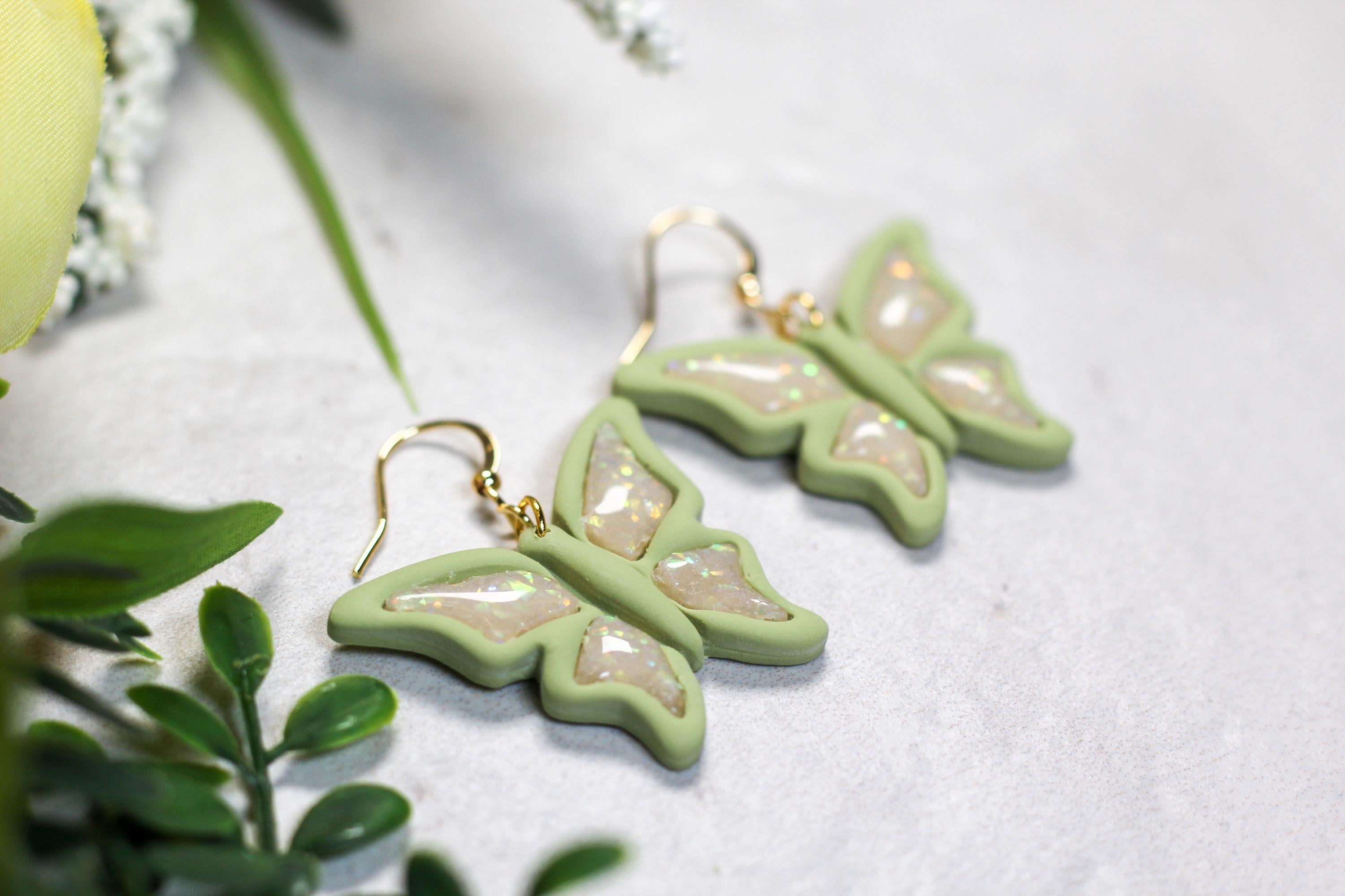 Green butterfly earrings on a white surface with green leaves in the background