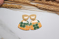 Pair of colorful earrings on a light surface with wheat stalks in the background