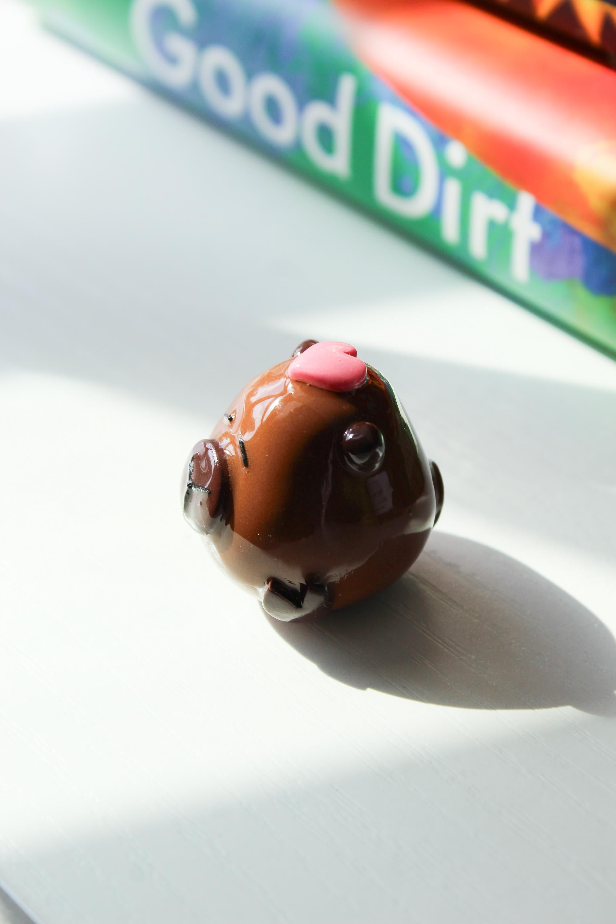 Small figurine of a Capybara with a pink heart on its head, placed on a surface with a book in the background.
