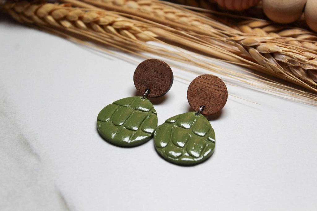 Green turtle shell earrings with wooden accents on a white surface with wheat stalks in the background.