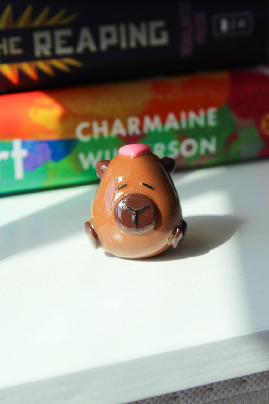 Small figurine of a Capybara with a pink heart on its head, placed on a surface with a book in the background.