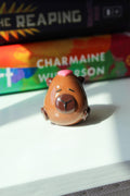 Small figurine of a Capybara with a pink heart on its head, placed on a surface with a book in the background.