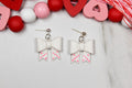 White bow-shaped earrings with pink heart details on a light gray background with decorative hearts and beads.