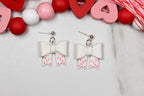 White bow-shaped earrings with pink heart details on a light gray background with decorative hearts and beads.