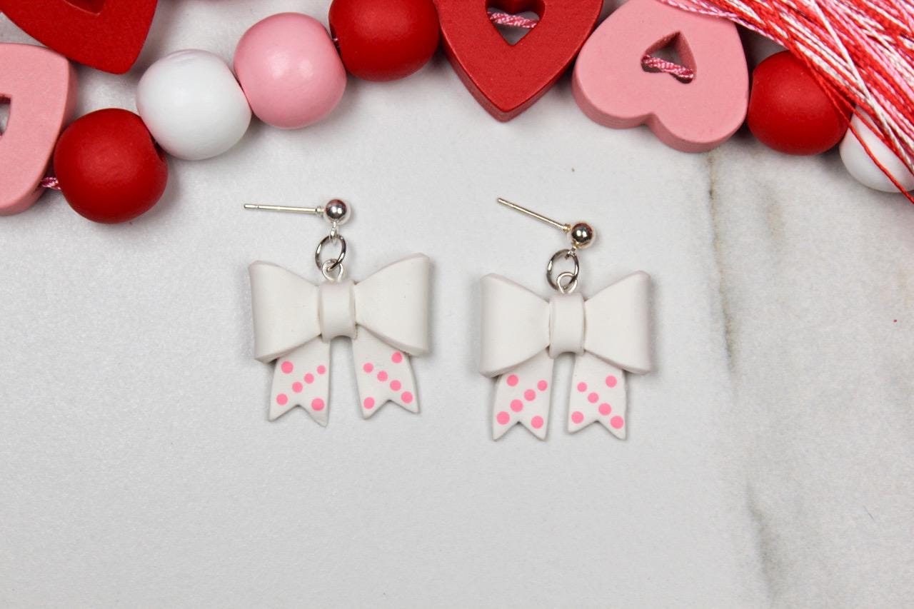 White bow-shaped earrings with pink heart details on a light gray background with decorative hearts and beads.