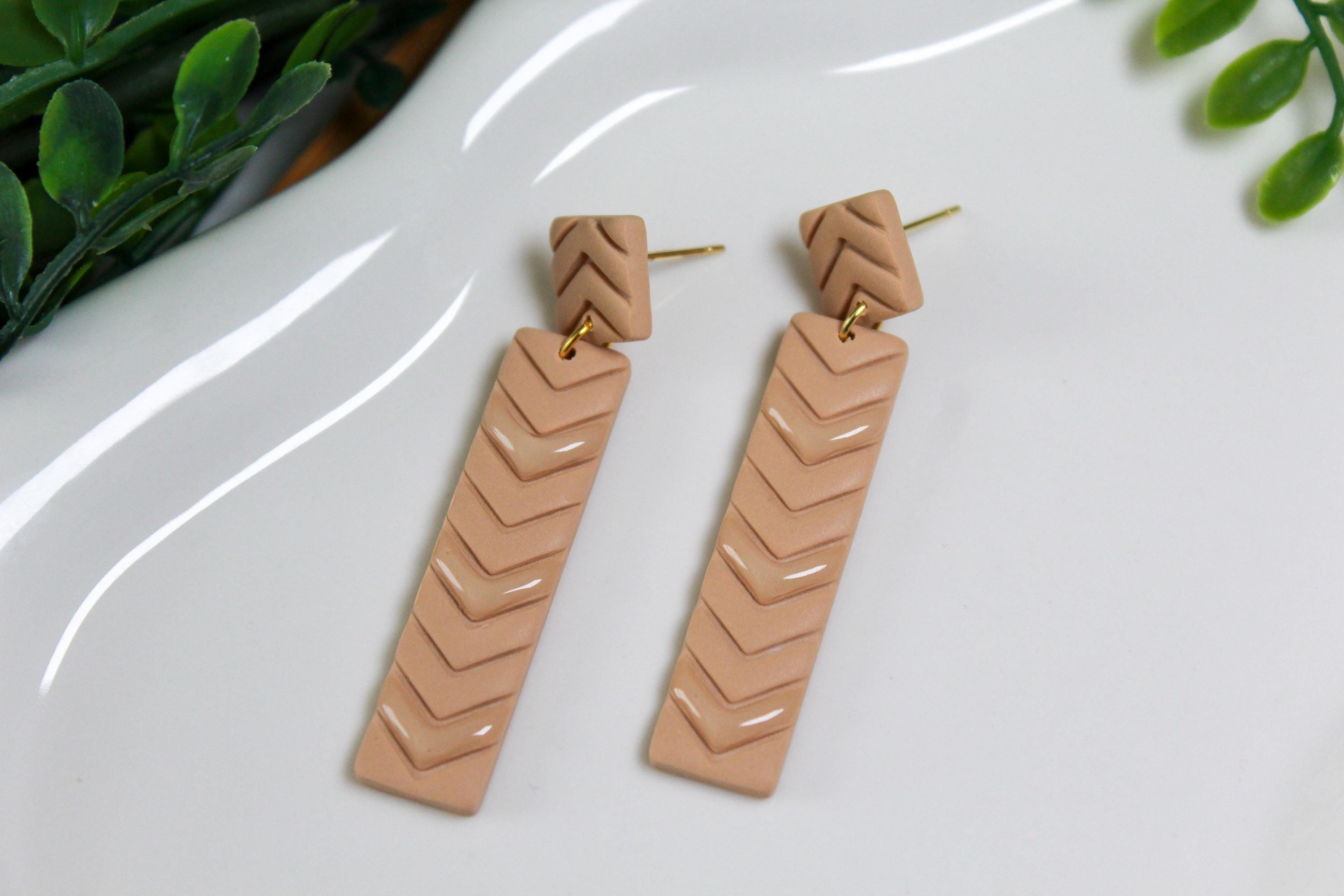 Pair of beige chevron patterned earrings on a white surface with greenery in the background.