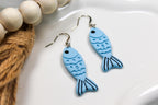 Blue fish-shaped earrings on a white surface with beads and twine.