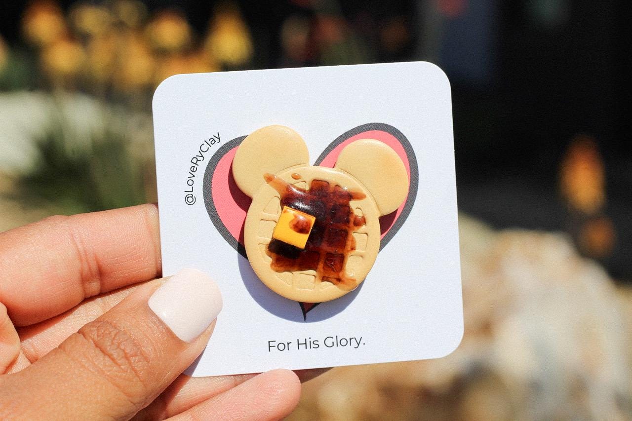Hand holding a card with a waffle-shaped pin featuring a heart and text, against a blurred natural background.