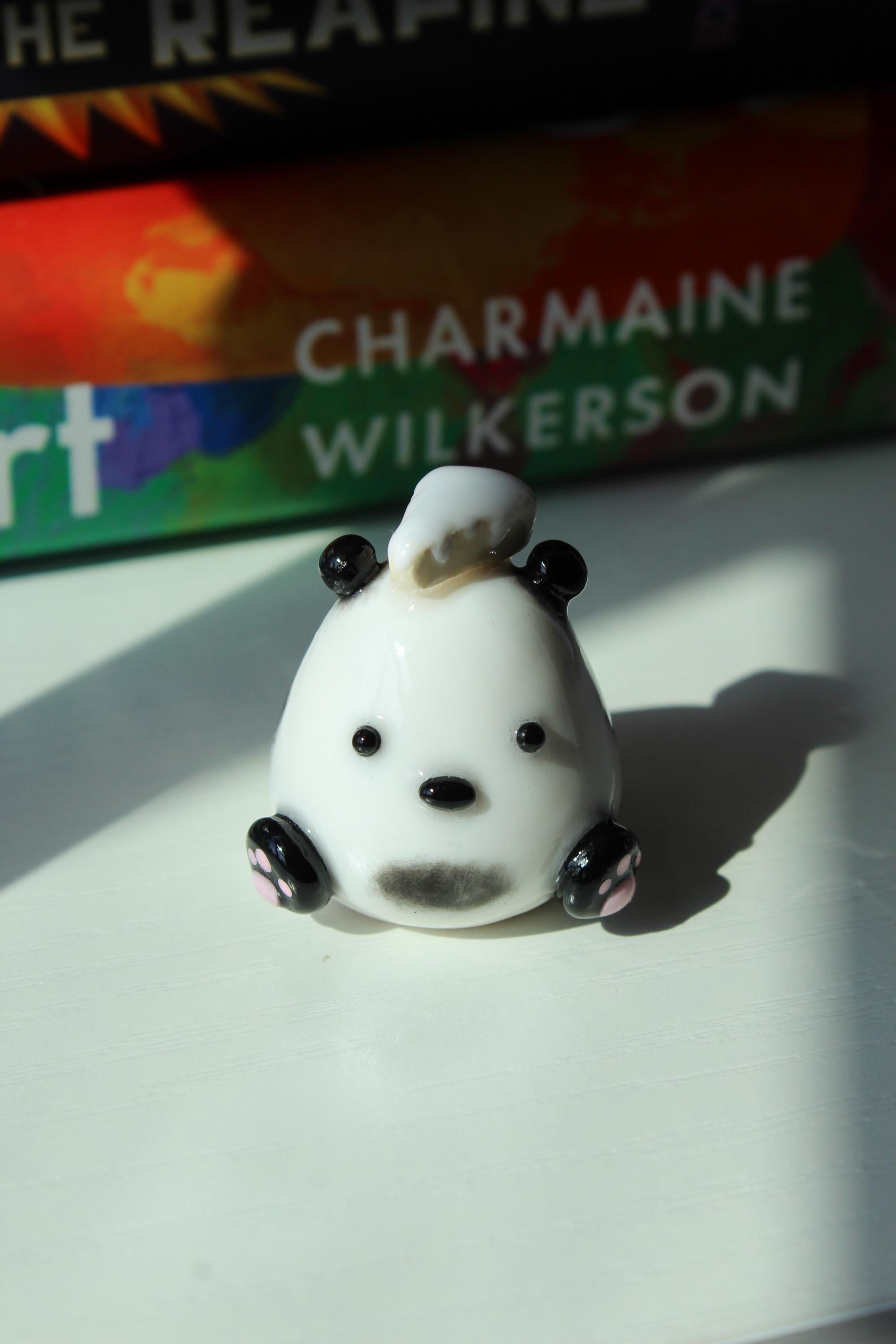 Small white figurine of a Panda with black accents and a piece of cake on a white surface, with a book titled 'Good Dirt' by Charmaine Wilkerson in the background.