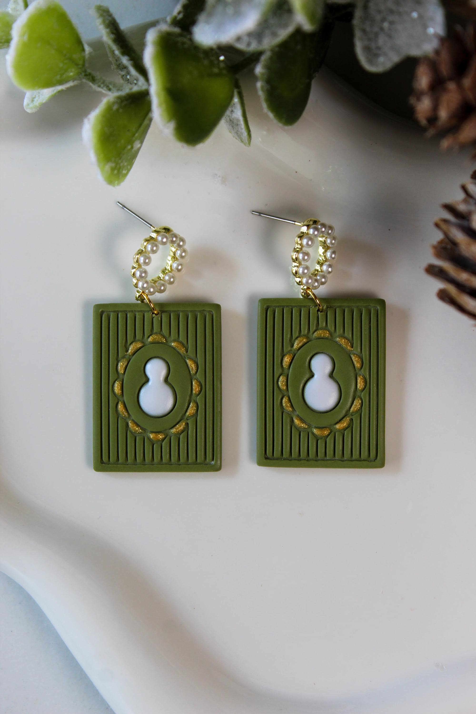 Green earrings with gold accents and snowman on a white surface with green leaves and pinecones in the background