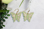 Butterfly-shaped earrings on a light surface with green leaves in the background