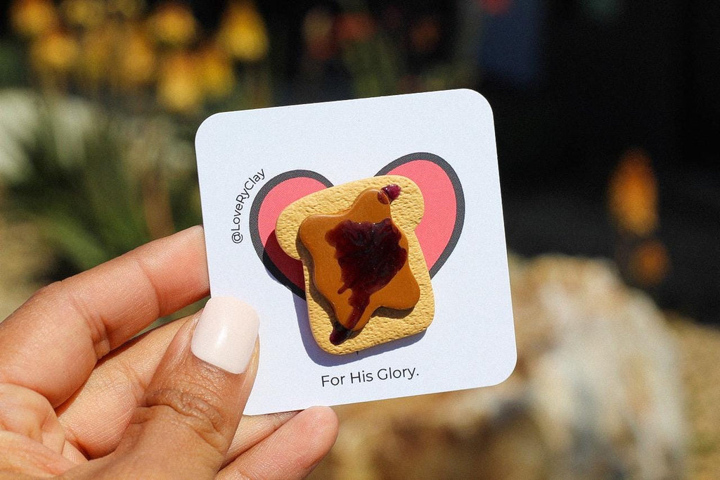 Hand holding a card with a heart-shaped design of a piece of bread with jelly, labeled 'For His Glory'.