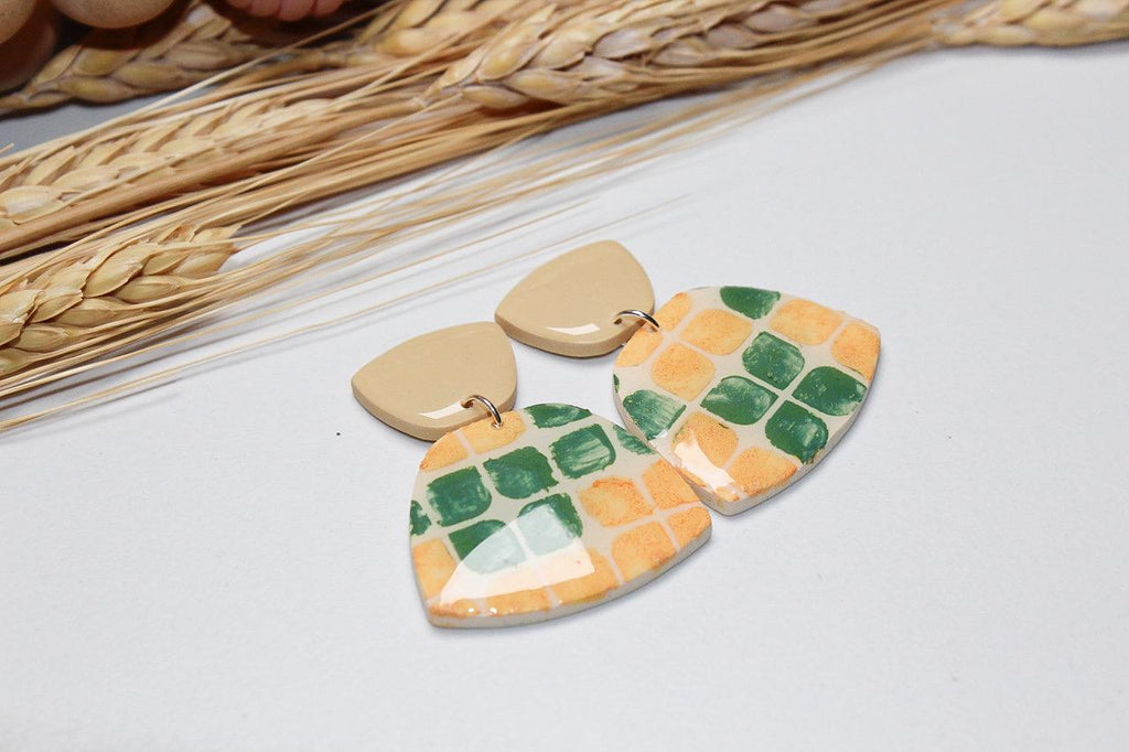 Pair of colorful earrings on a light surface with dried wheat stalks in the background